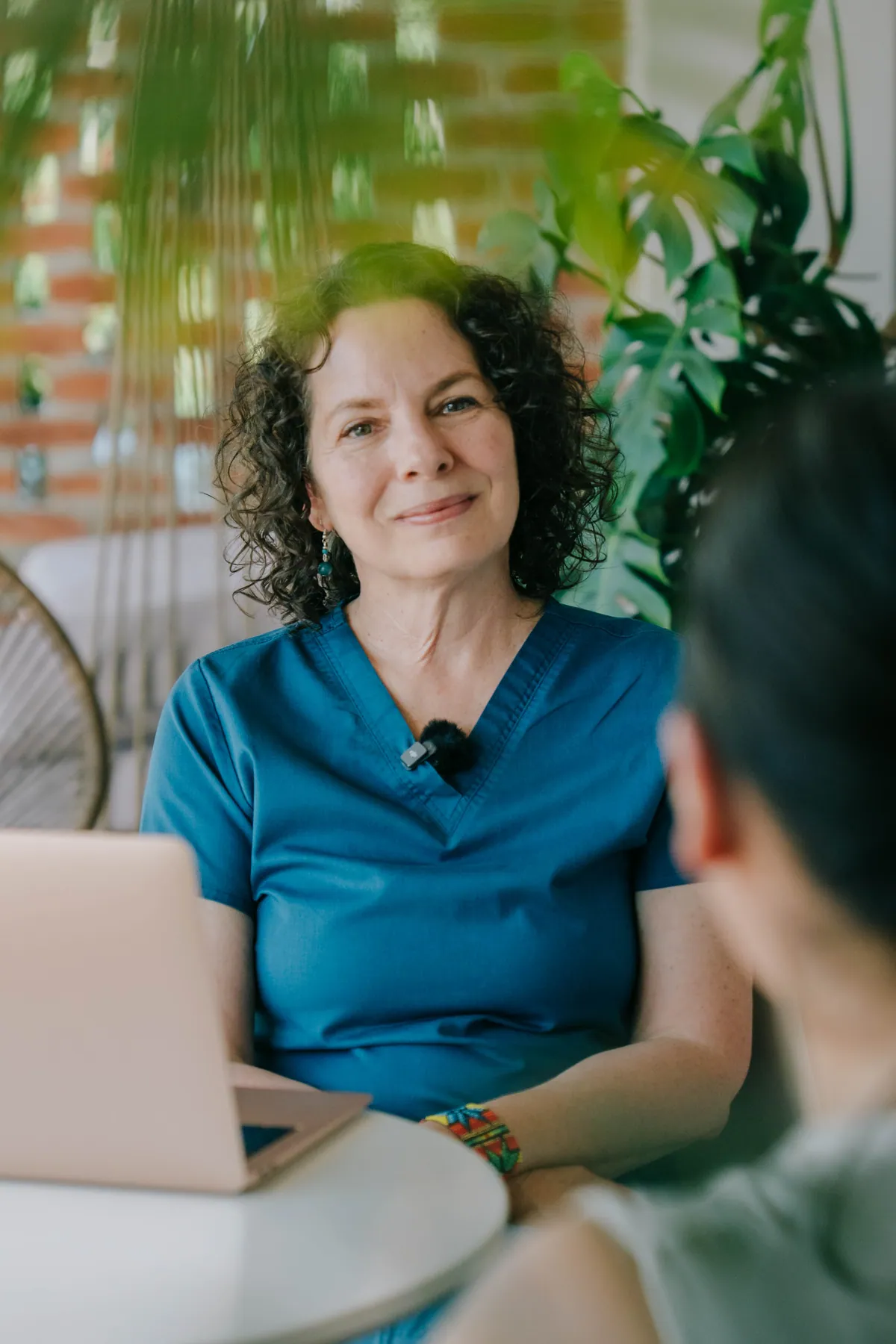 The lodge doctor in blue scrubs at a small white table with a laptop, smiling at a guest seen from behind, with plants and a brick wall behind her