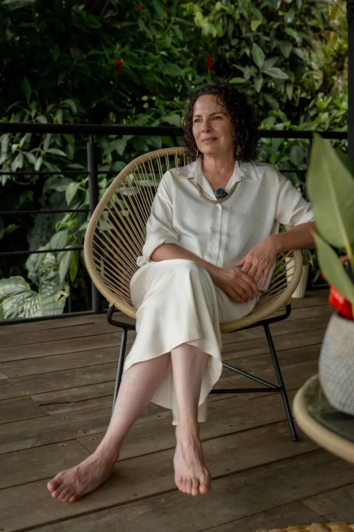 The lodge doctor seated barefoot in a woven chair on a wooden terrace, in a cream shirt and skirt, dense green leaves behind her