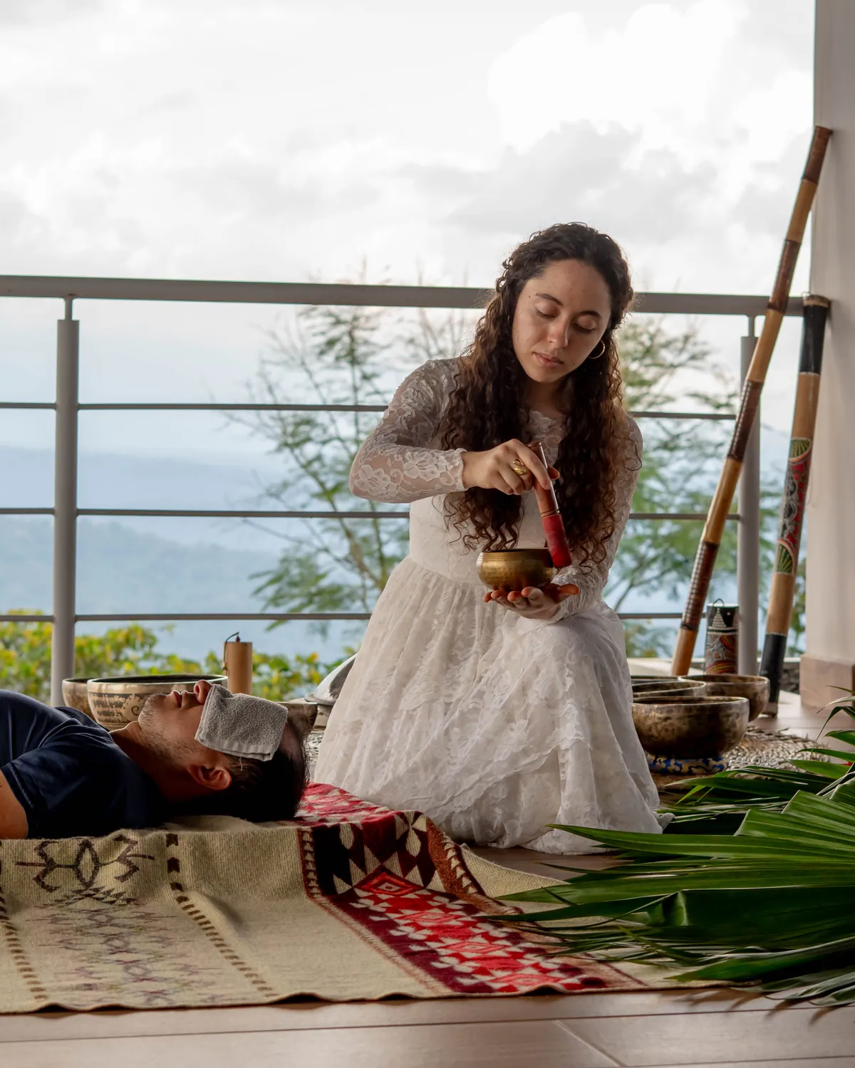 A woman in a white lace dress kneeling on a terrace above the valley, sounding a brass singing bowl over a guest lying on woven rugs