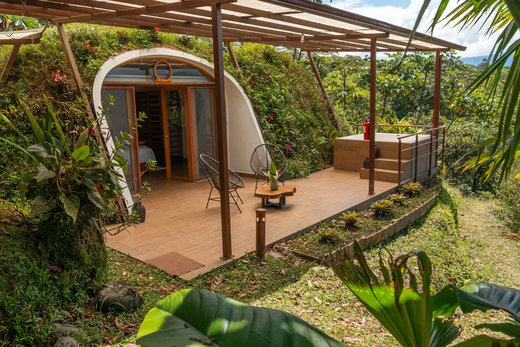A jungle dome with an arched glass front under a wooden pergola, two chairs on its deck and a hillside of plants around it