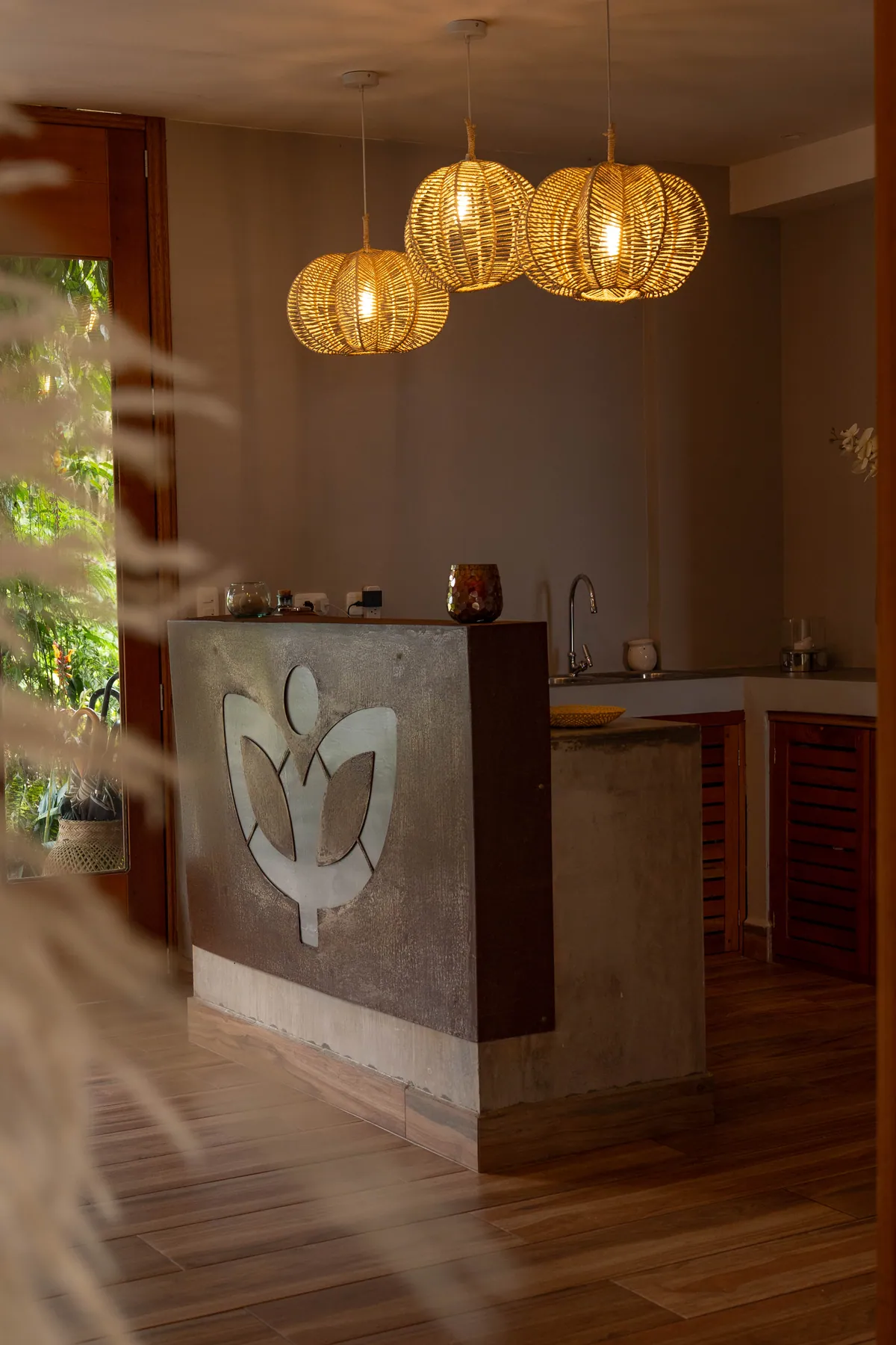 The lodge reception at dusk: three woven pendant lamps over a concrete counter carrying the Vibra sprout mark, a doorway to the garden beyond
