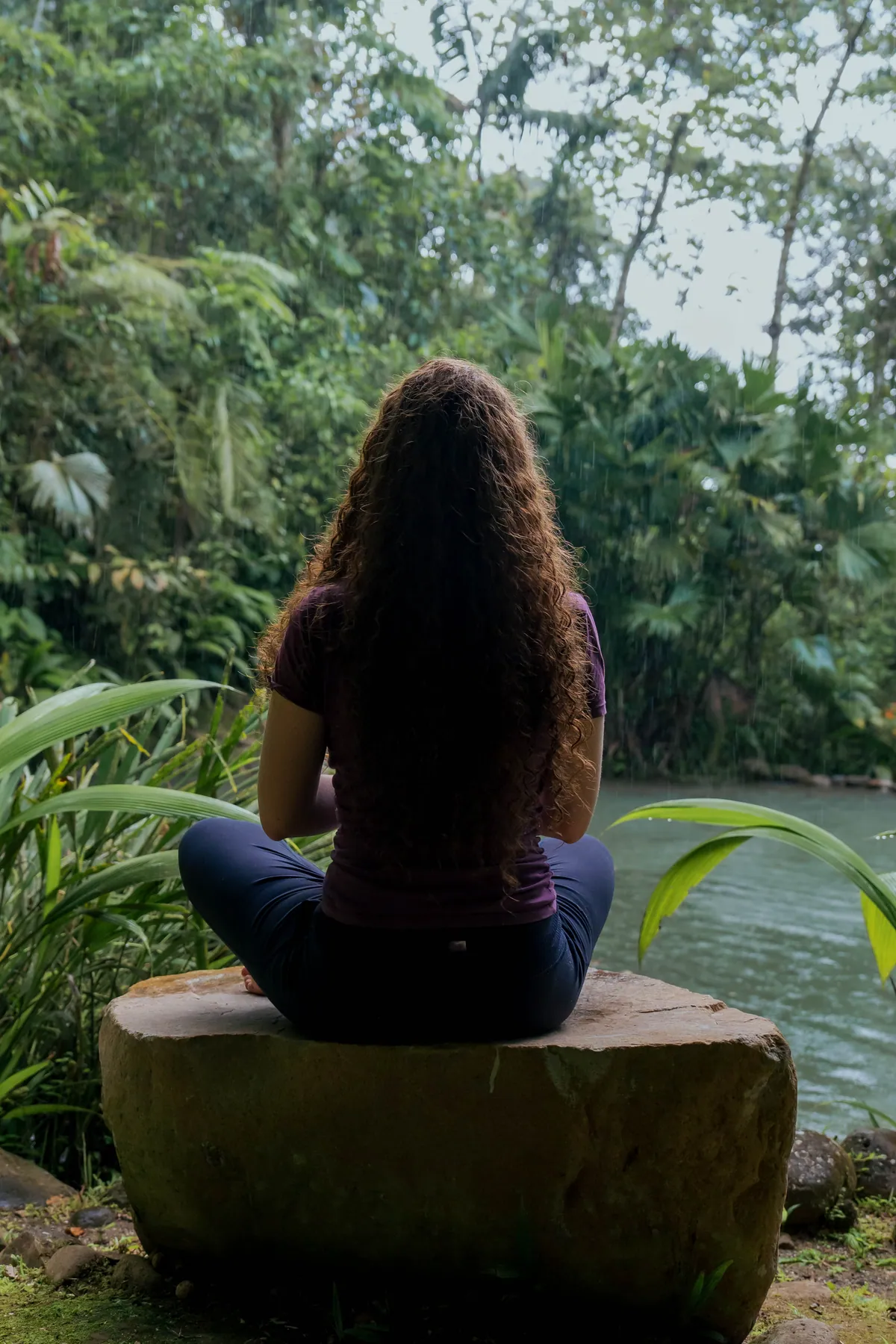 A woman sitting cross-legged on a flat stone above a green river, her back to the camera, rain falling through the palms
