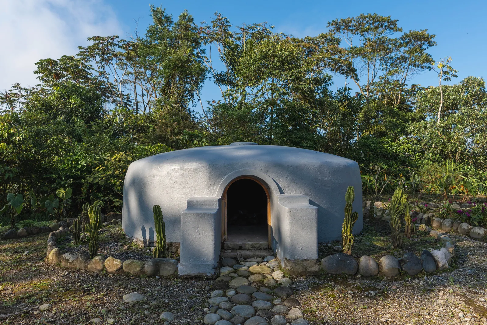 A low domed temazcal of pale rendered stone with an arched doorway, ringed by river stones and cacti under the forest canopy