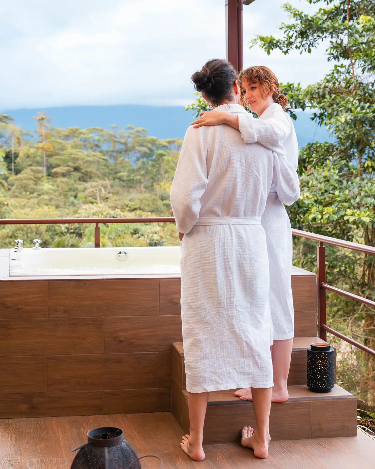 Two guests in white robes on a wooden terrace beside a filled outdoor tub, the forested valley behind them
