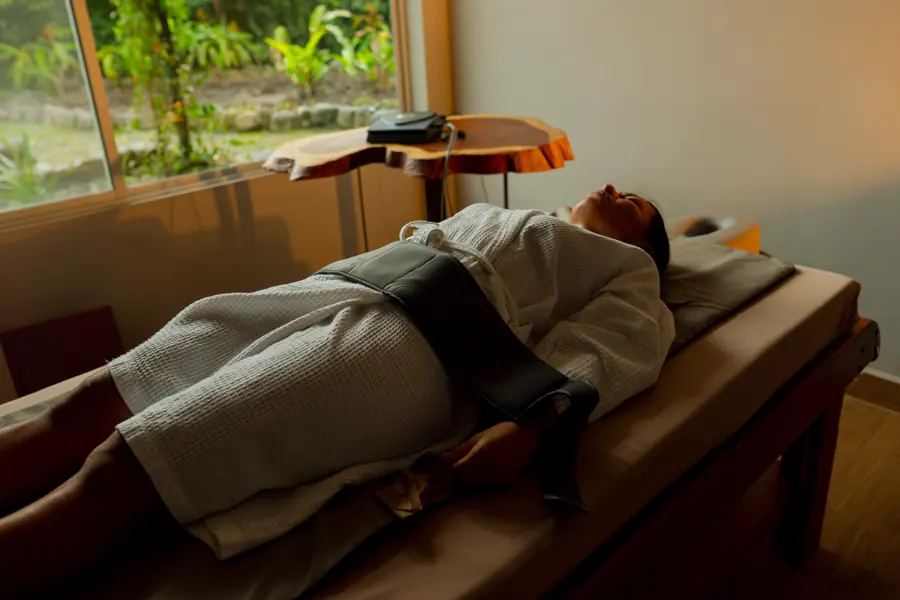 A guest resting under a blanket on a treatment table, beside a window onto the forest