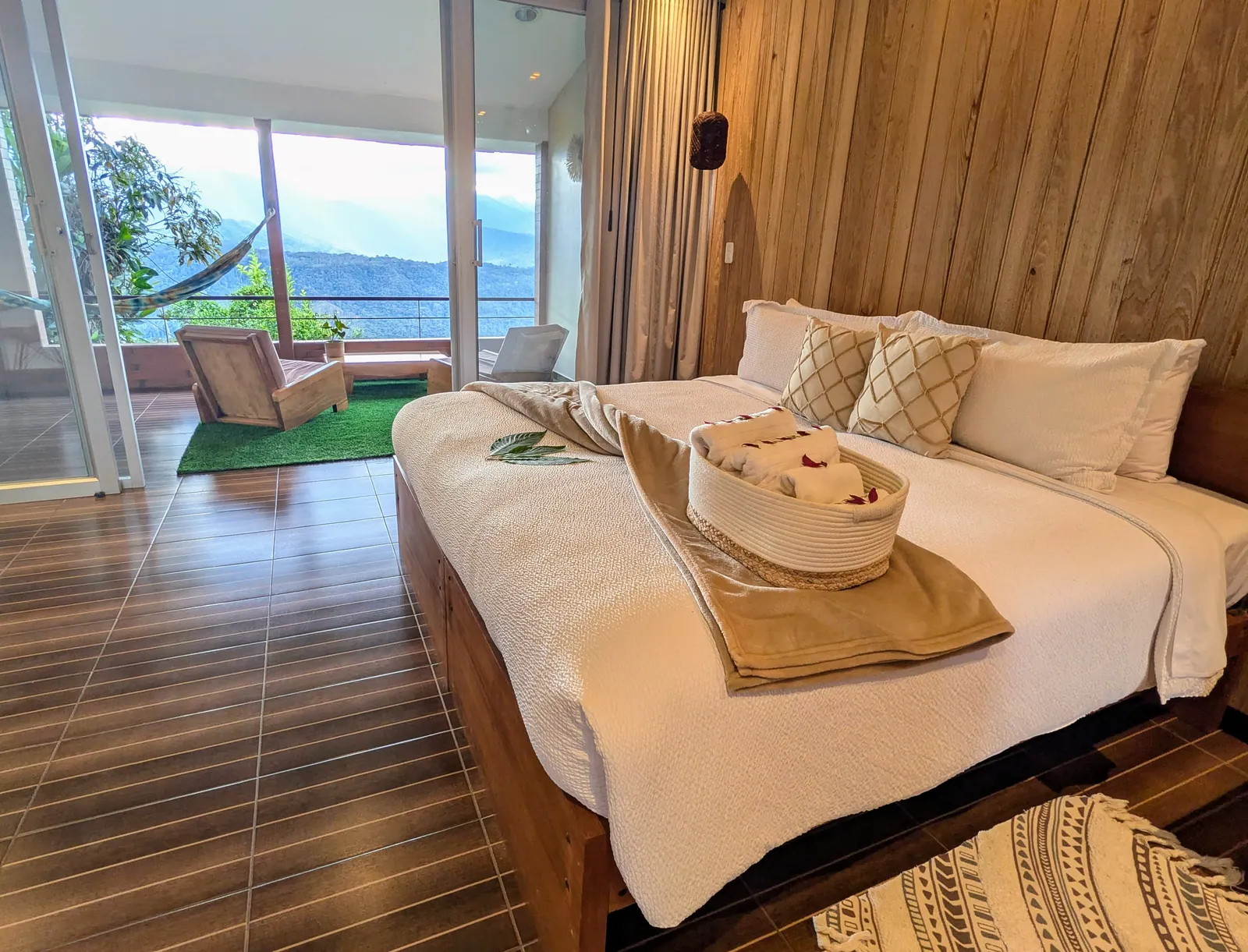 A bed against a wood-panelled wall, sliding doors open onto a terrace with a hammock and the valley beyond