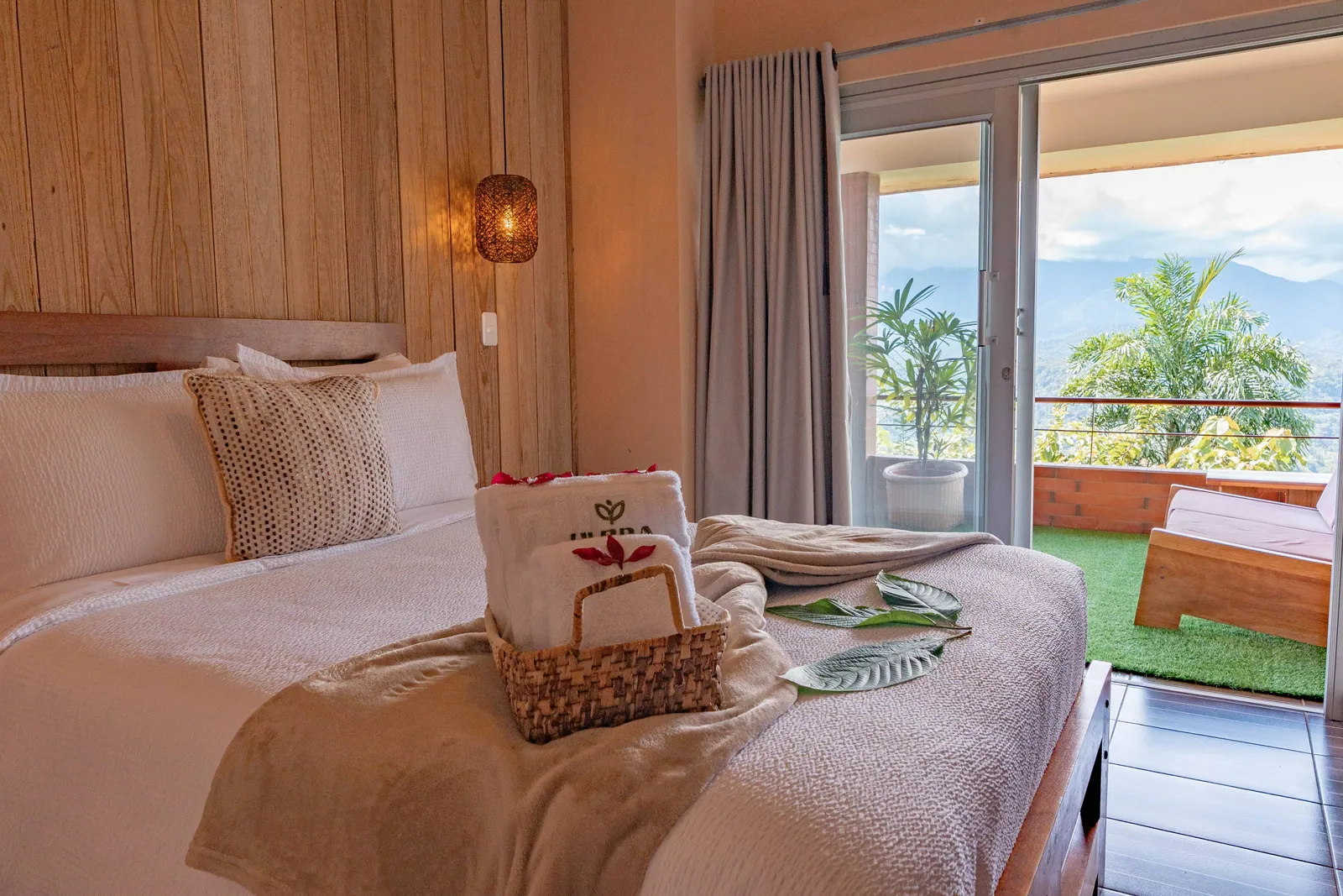 A bed with a basket of towels and leaves on the cover, a curtain drawn back from an open door onto a terrace facing the mountains