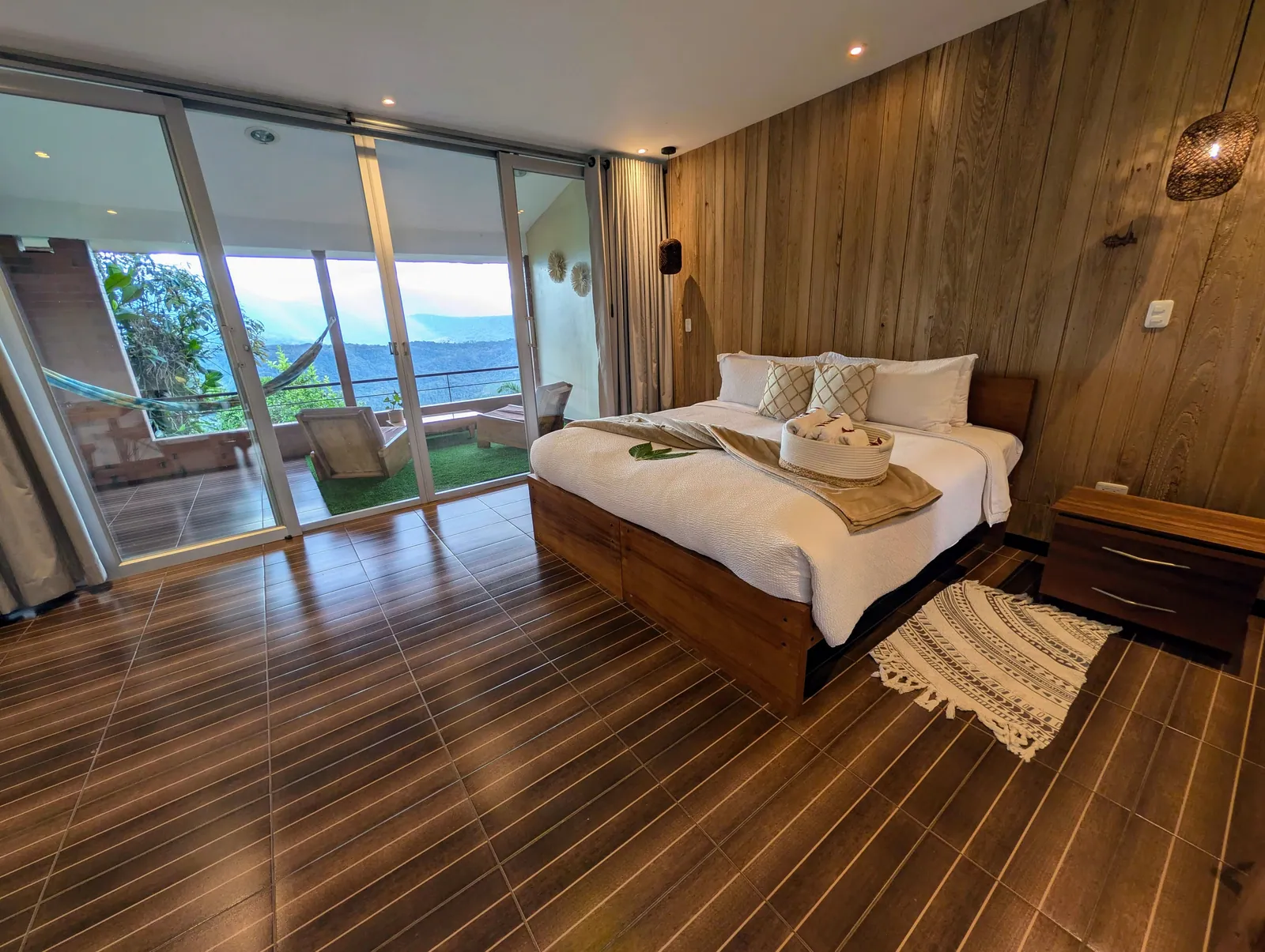 A room seen from its doorway: a bed, a wood-panelled wall, and the terrace with its hammock behind the glass