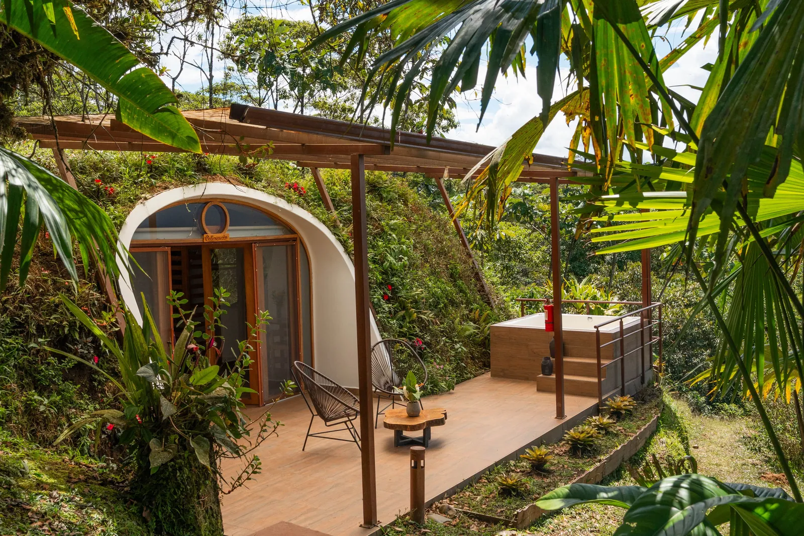 A jungle dome with an arched glass front, two chairs and a wooden tub on its covered terrace, palms and ferns around it