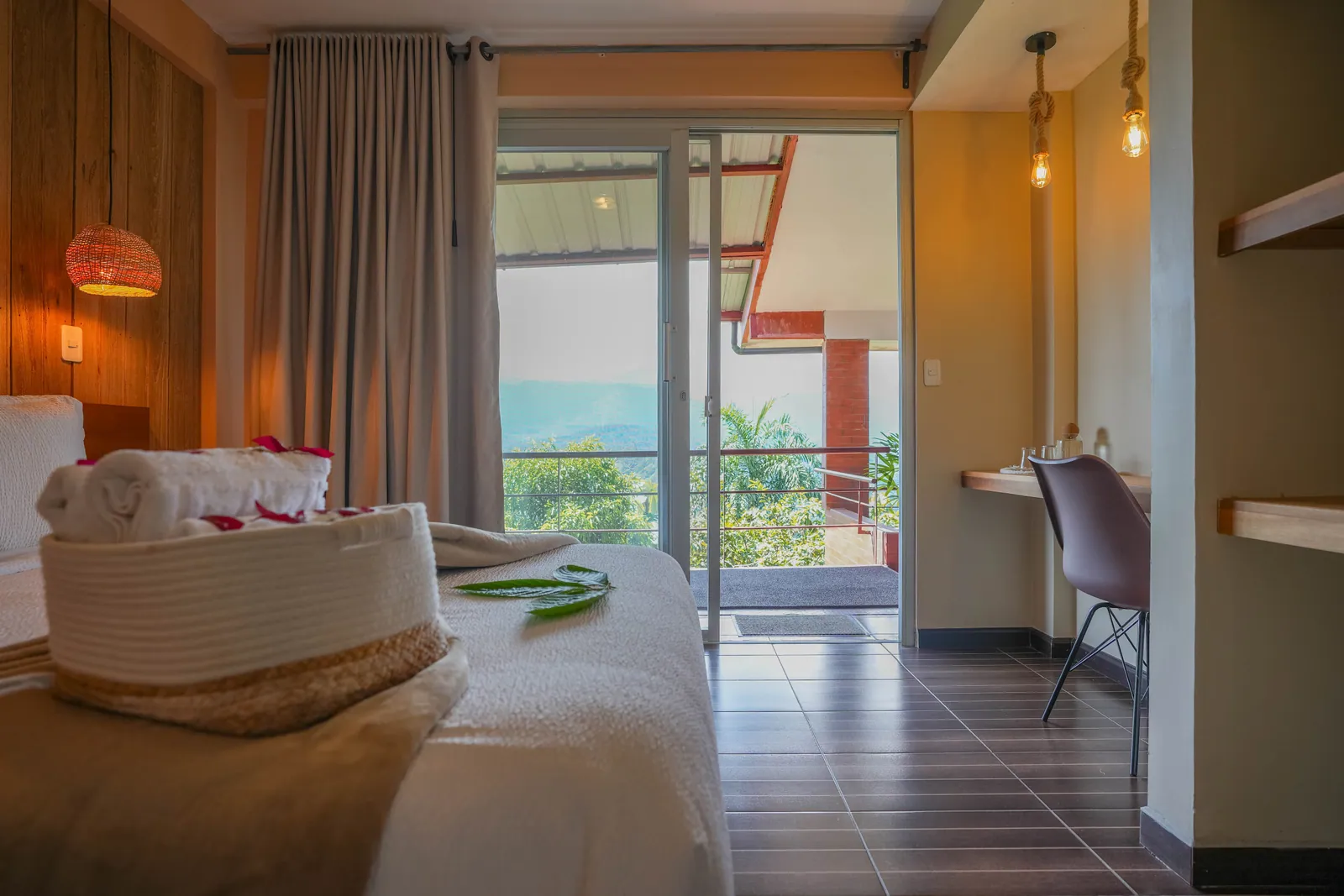 A room with a woven hanging lamp, a bed with towels laid on it, and a desk beside the open door to the terrace